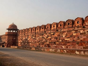 Agra Fort's red sandstone ramparts seen from across a dusty road at late afternoon, long shadows across the foreground, fort wall filling the upper two-thirds of the frame, overcast warm sky above