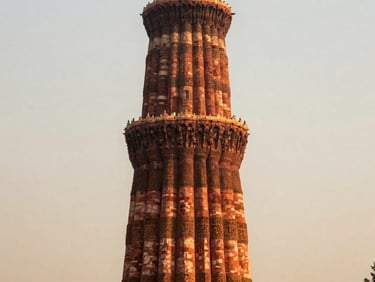 Close framing of the Qutb Minar at dawn, warm sidelight catching the red sandstone fluting, sky pale gold behind the tower, Delhi's haze soft in the background