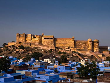 Panoramic view of Mehrangarh Fort rising above Jodhpur's blue rooftops at dusk, golden sidelight on the sandstone battlements, hazy indigo sky above, vast landscape scale