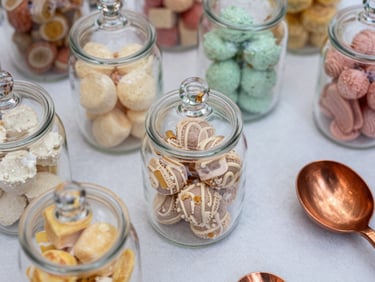 Extreme close-up overhead of a styled candy table surface: glass apothecary jars filled with pastel confections, copper scoops resting on white linen, bright natural daylight, shallow depth of field revealing jar texture and ribbon detail