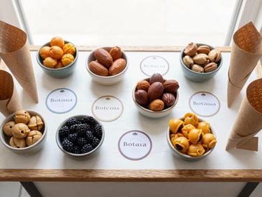 Overhead flat lay of a styled snack cart top surface: small ceramic bowls of botanas arranged by color and size, kraft paper cones, coordinated label tags, bright overcast natural daylight, white and warm-wood surface