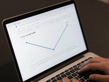 Close-up overhead of a laptop screen displaying a Google Ads dashboard with declining cost-per-lead metrics, a hand resting near the keyboard, neutral studio light, dark background