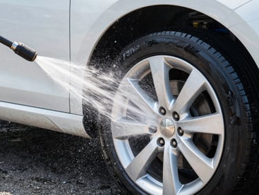 Crisp commercial photography of a high-pressure water jet atomizing over a clean car wheel, bright natural daylight, water droplets frozen in mid-air, sora-blue reflections