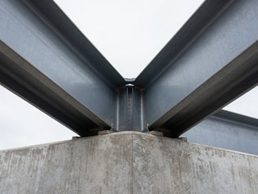 Symmetrical close-up of heavy structural steel beams meeting a site-poured concrete foundation, high-contrast overcast daylight emphasizing the raw metallic and concrete textures.