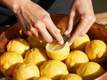 Hands carefully zesting fresh organic lemons over a copper vessel, warm sun-drenched light, sharp shadows, editorial photography.