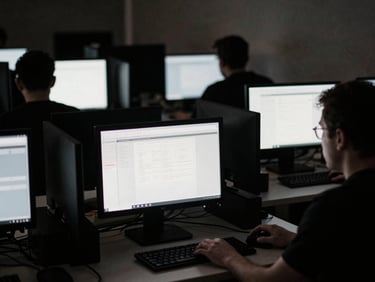 Low-key shot of multiple computer monitors glowing in a dark room, silhouettes of operators, gritty film grain