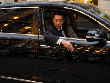 Cinematic portrait shot of a professional chauffeur in a dark tailored suit opening the door of a pristine black executive sedan, soft golden reflections of city lights on the polished car body, 35mm lens.