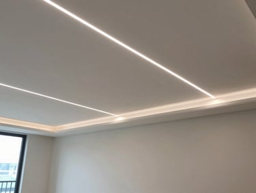 Wide environmental shot of completed commercial interior ceiling with continuous recessed LED linear channels integrated into drywall coves, indirect light washing a plastered wall, single architectural vantage point, controlled daylight from left, no people