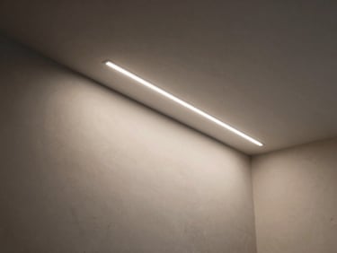 Architectural detail of recessed LED linear profile installed in a drywall ceiling cove, soft indirect light spilling downward on a finished plaster wall, north-facing daylight supplement, precise installation geometry visible