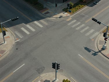 Overhead aerial view of a urban road intersection with traffic signal infrastructure visible at each corner, roads extending to the frame edges, neutral midday light, no vehicles as the primary subject