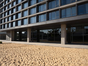 modern architectural photography, sharp natural sunlight, deep shadows, warm sand and charcoal tones, wide-angle lens, 35mm, sleek commercial glass and concrete building exterior