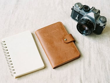 Flat-lay of a neatly arranged leather passport holder, notebook, and vintage camera on a cream linen sheet, soft overcast daylight, 35mm film aesthetic.
