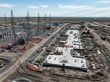 Aerial drone view of a graded industrial land parcel, massive concrete foundations, high-voltage substation under harsh midday sun, high-contrast slate and utility orange tones