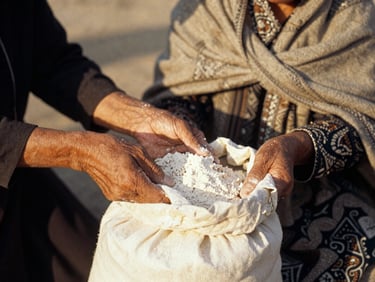 Close-up of hands distributing a sack of flour to a widow wearing a traditional shawl, warm golden-hour sunlight, dignified documentary style, 35mm film.