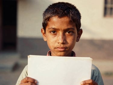 A close-up portrait of a young Pakistani child holding a clean schoolbook, looking directly at the camera with agency, warm natural daylight, shallow depth of field, 35mm film style.