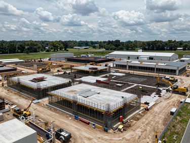 Wide aerial of an active commercial construction site, steel framing and concrete slabs mid-pour, equipment visible, overcast Tennessee sky, midday natural light