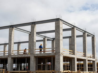 Eye-level wide shot of a commercial building frame under construction in natural daylight — exposed concrete columns and steel beams forming a multi-storey grid, construction crew visible at mid-distance focused on structural work, overcast sky above