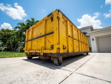 Low-angle heroic shot of a bright yellow steel roll-off dumpster on a clean concrete driveway, harsh South Florida midday sun, high contrast.