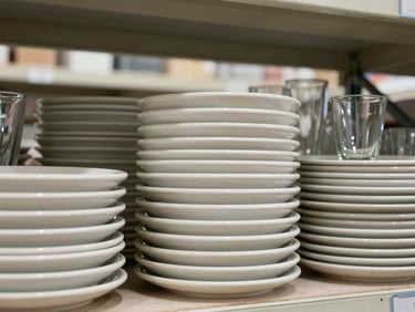 Eye-level shot of stacked ceramic plates and glassware arranged on warehouse shelving, flat natural light from above, no people, neutral background, clear texture of stoneware