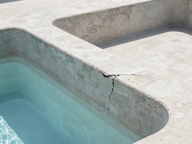 Close-up of a deteriorated concrete pool shell with visible structural cracking, stained plaster, and empty basin, harsh daylight.