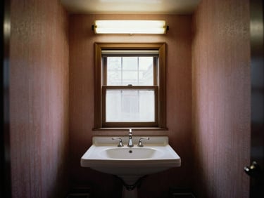 Straight-on architectural shot of a dated 1980s master bathroom, dark cramped layout, dusty pink wallpaper, harsh fluorescent lighting.
