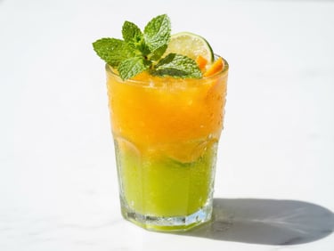 Macro close-up of a condensation-covered glass filled with a vibrant orange and lime layered juice, fresh mint leaves on top, bright natural sunlight casting crisp shadows on a clean white background