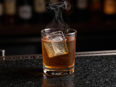 Low-key dramatic close-up of a hand-poured cocktail on a dark basalt counter, single warm key light catching the amber liquid and rising vapor, dark background.