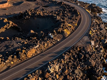 Crisp top-down aerial drone photograph of a coastal highway winding through dark volcanic rocks, golden hour light highlighting the asphalt texture.