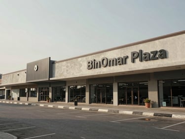 Wide environmental daytime shot of BinOmar Plaza exterior, full facade of street-level retail storefronts stretching left to right, natural overcast light, concrete and masonry materiality, parking approach visible in foreground, no people staged, documentary framing