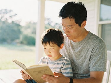 Warm editorial photography of a father and young son reading together on a sunlit porch, soft morning daylight, shallow depth of field, 35mm film style.