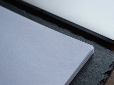 Close-up of heavy textured paper documents resting on a dark slate desk, architectural shadows, cool overcast daylight.