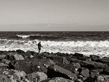 High-contrast monochrome photograph of a lone fisherman on a rocky Ordu shore, big dramatic dark waves, overcast sky, fine art photography