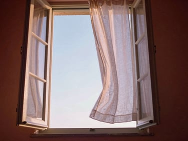An open window looking out to a soft blue sky, light linen curtain blowing in the wind.