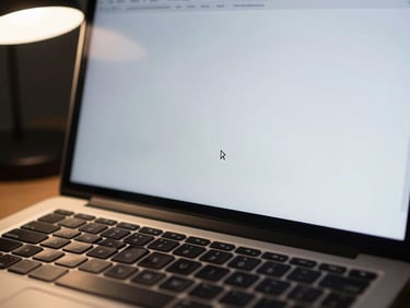 Laptop screen showing a blank document with cursor blinking, keyboard in foreground, cool desk lamp light, no faces, tight crop on screen edge and keys