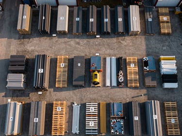 Bird's-eye overhead view of an organized job-site material staging area, pallets sorted by trade — roofing sheets, plumbing fittings, electrical conduit — each labeled with phase markers, golden-hour exterior light casting long shadows across the organized grid
