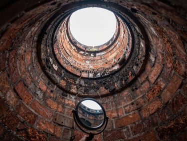 Wide angle looking up inside a brick chimney flue, technician's inspection mirror and light tool visible at the bottom edge, soot deposits on the flue walls, natural daylight at the top opening