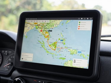 Close-up of a logistics route planning map on a tablet screen mounted in a truck cab, showing Java island with highlighted distribution corridors and waypoints, dashboard instruments visible at the bottom edge, natural light from the windshield