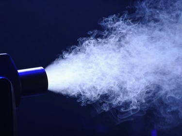 Close-up of a professional stage fog machine nozzle emitting dense white smoke under high-contrast blue spotlighting, technical detail