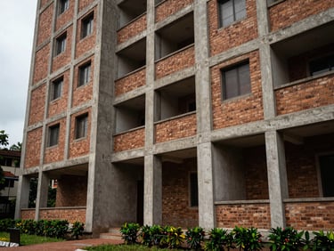 Ground-level exterior of a mid-rise apartment building in Perumbakkam, Chennai, daylight overcast, raw concrete columns and brick facade visible, modest landscaping in foreground