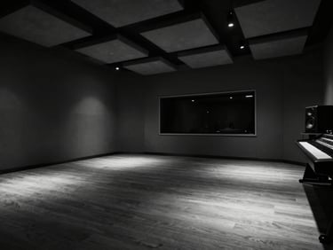 Wide architectural shot of an empty recording studio interior, deep shadows pooling in the far corners, a single studio monitor glowing faintly at the right edge, geometric ceiling panels receding into darkness, high-contrast monochrome atmosphere, extreme negative space dominating the frame