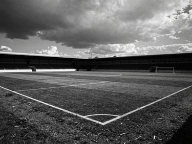 Editorial black and white photography, dramatic natural light, deep shadows, high contrast, 35mm film grain. An empty regional football pitch in Seville under a heavy overcast sky, dramatic dark clouds, white chalk lines on the dark soil.