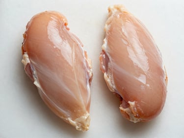 Close-up overhead of two raw chicken breast fillets on a clean white cutting board under flat studio light, pale surface showing natural muscle lines and clean trim, no props, no seasoning, tight frame filling the full width