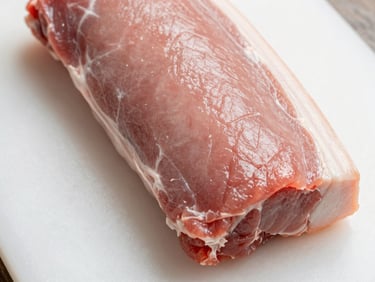 Close-up overhead of a raw pork loin section on a clean white cutting board under flat studio light, pale pink flesh with a uniform fat cap, no props or styling, tight frame showing surface texture and clean cut edge