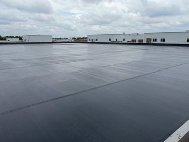 Wide aerial view of a flat commercial rooftop membrane installation, overcast daylight, HVAC units visible in background, clean seam lines in foreground, no people, honest documentary framing