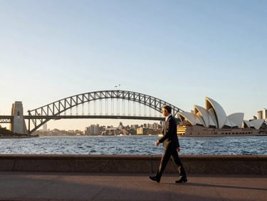 Wide panoramic shot of Sydney Harbour at golden-hour morning light, the bridge and opera house visible in the mid-distance, a professional walking along the waterfront promenade in the foreground, the scale of the city readable behind them