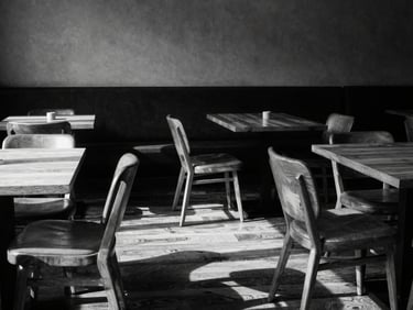 Wide-angle shot of a minimalist café seating area, empty chairs, warm wood tables, strong shadows, high-contrast monochrome tones, 35mm lens.