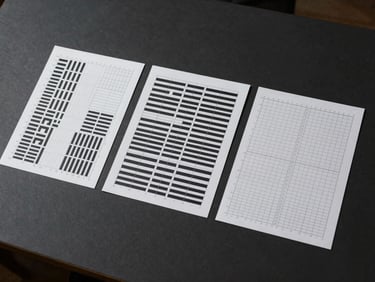 Overhead studio shot of three printed design grid templates arranged on a matte dark surface, each sheet showing typographic hierarchy systems with visible baseline grids and column markers, diffused studio light from above, no hands or people