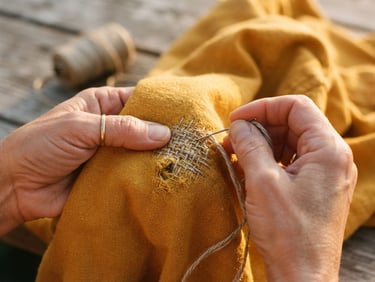 Close-up of hands carefully mending a heavyweight marigold organic cotton garment with thick linen thread, warm golden-hour sunlight casting soft shadows, 35mm film look.