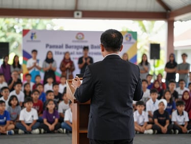 Medium shot from behind the MC standing at a podium on an open stage at a children's competition event in Yogyakarta — large audience of children and parents visible ahead, banners and competition signage in background, natural daylight from open hall windows, MC gesturing toward participants