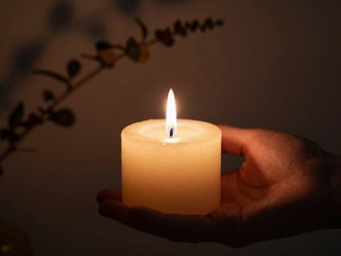 Close-up of a hand gently holding a single burning candle, soft focus on the flame, deep shadows, warm chiaroscuro lighting, botanical sprig in the background.